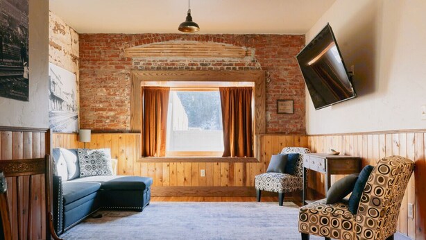 A cozy room with a brick wall, a large window with curtains, a blue sofa, patterned chairs, a wooden floor, and a flat-screen TV mounted on the wall. - Nugget Suite - Main Street (Deadwood)