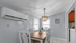 A dining area with a wooden table and white chairs, an air conditioning unit, and a chandelier. - The Teal Door is a 3 Bedroom 1 Bath Unit. Sleeps 6. Linens Included! (Carolina Beach)
