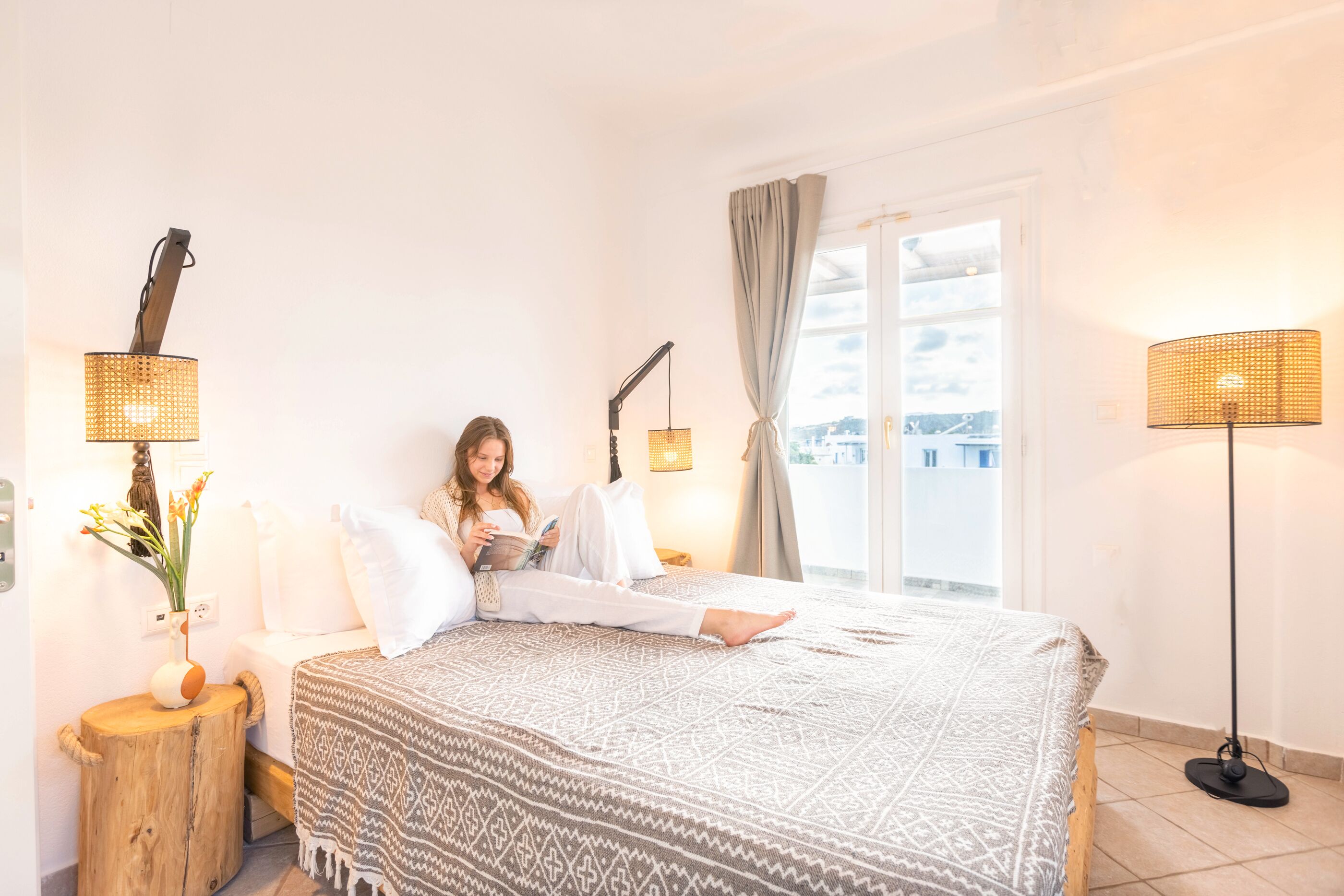 Un dormitorio con una cama, una mesita de noche de madera, una lámpara de pie y una ventana con cortinas.
