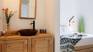 Une salle de bain avec un meuble-lavabo en bois, un grand miroir, un lavabo sur colonne et un lit recouvert d’une couverture à motifs.