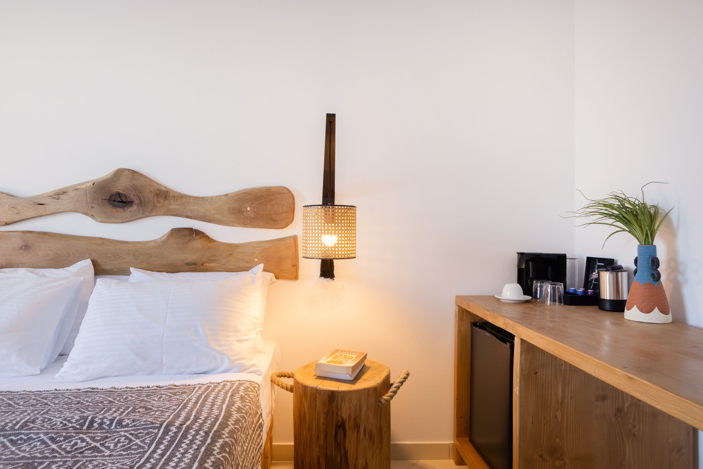 A bedroom with a wooden headboard, a bedside lamp, a small wooden stool with a book, a bedside table with a coffee maker, and a plant.