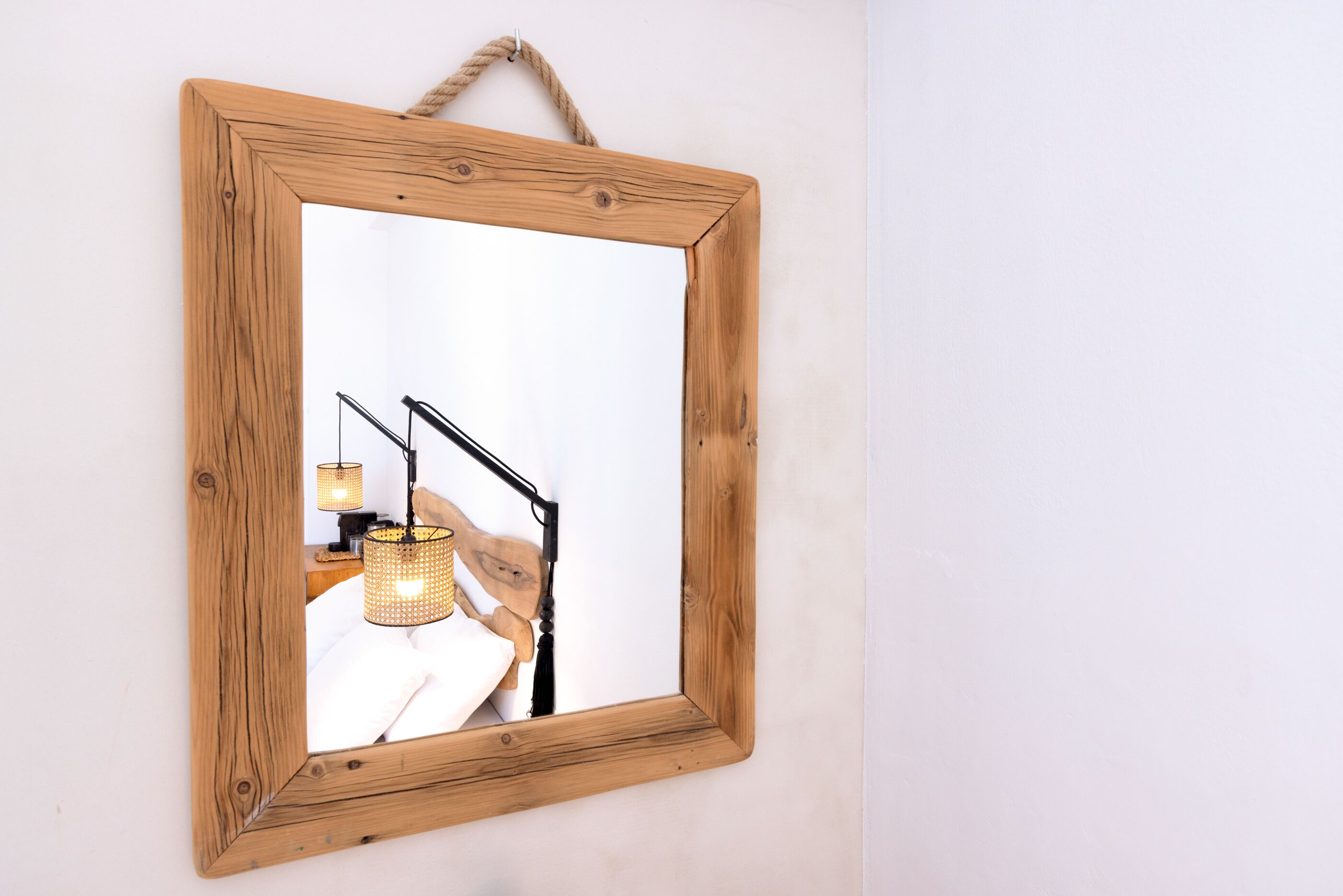 A wooden-framed mirror with a rope handle, reflecting a bed with a lamp and a hanging basket.