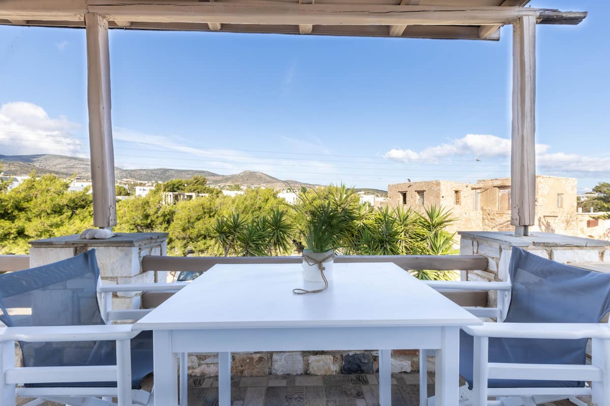 A white table with blue cushions under a pergola, overlooking a village with mountains in the background.