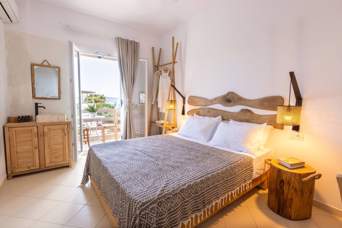 A bedroom with a bed, a wooden nightstand, a mirror, and a view of the outdoors.