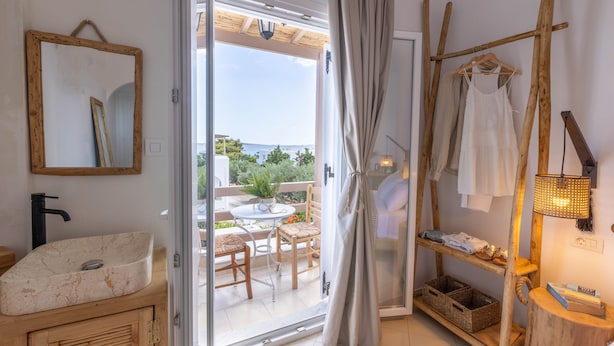 Une salle de bain avec un lavabo en marbre, des meubles en bois, un miroir et une vue sur une terrasse extérieure équipée d’une table et de chaises.