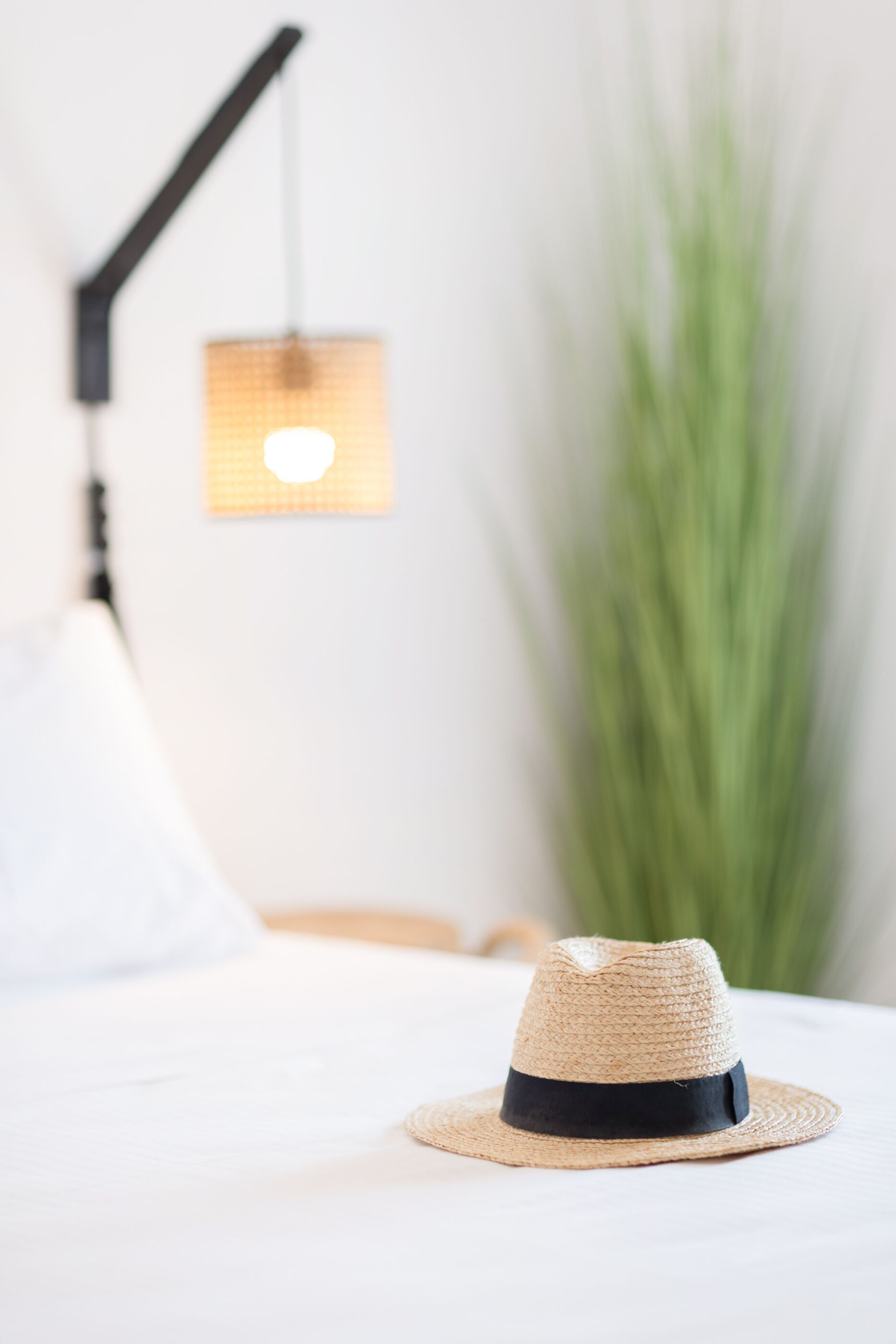 Un sombrero de paja sobre una cama, con una lámpara y una planta al fondo.