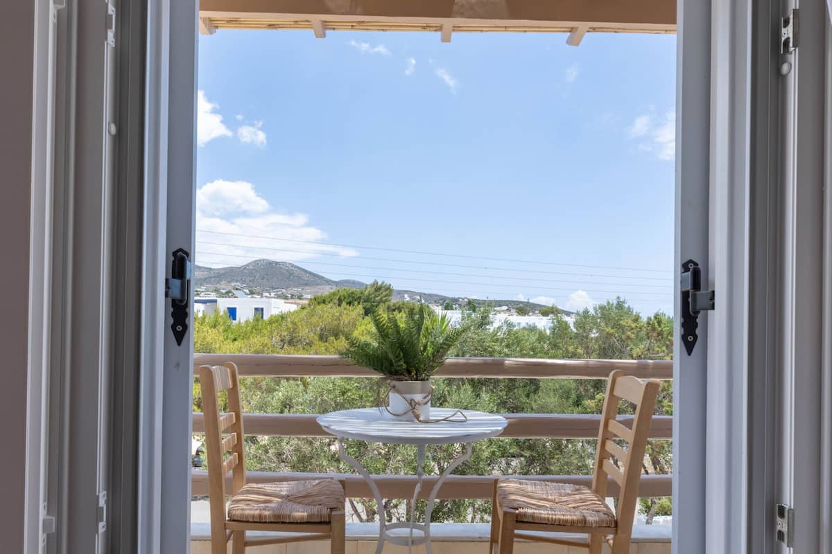 An open door leading to a balcony with two wooden chairs and a small table, overlooking a scenic landscape with greenery and hills.