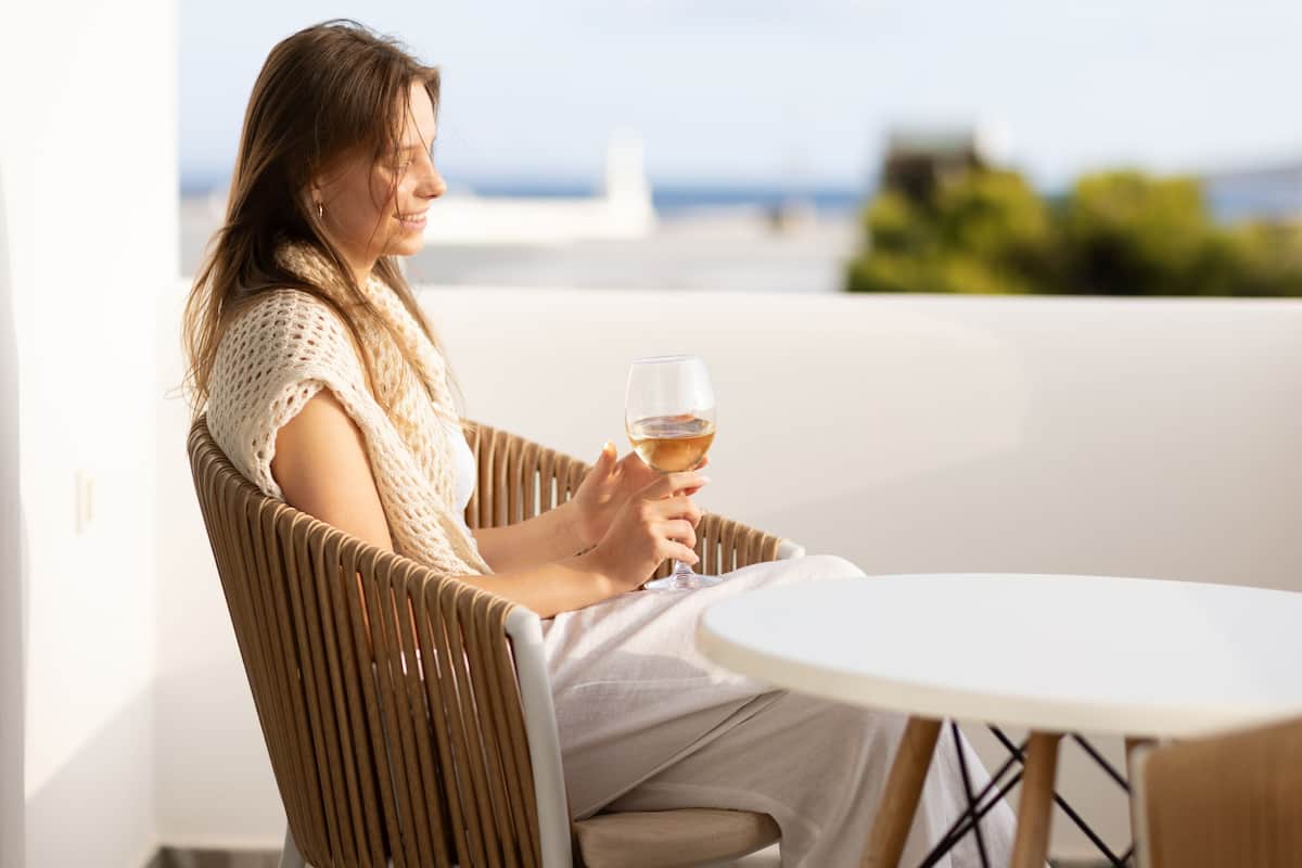 A person sitting on a wicker chair at a round table outdoors, holding a glass of wine.