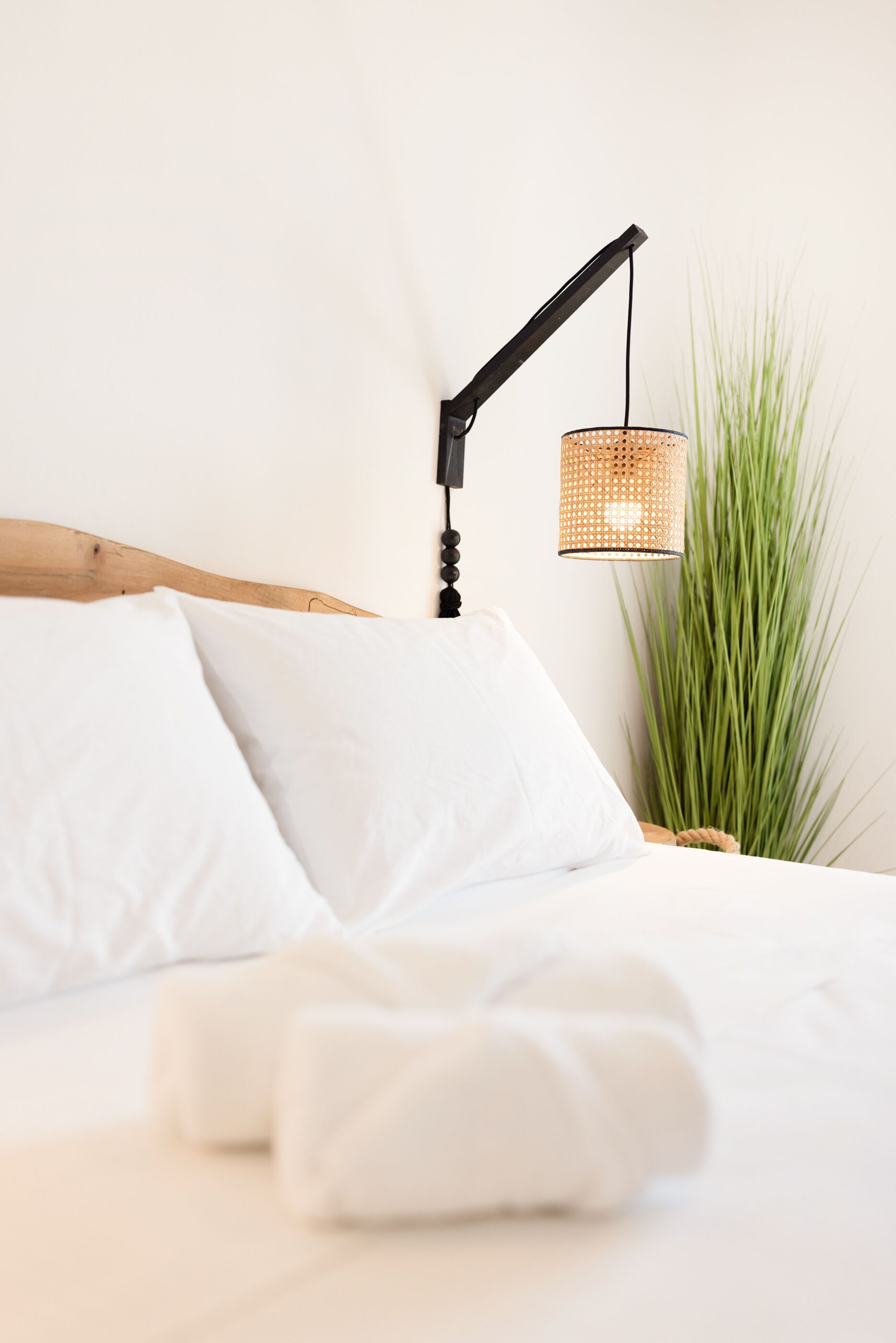 Ein ordentlich bezogenes Bett mit weißen Leinen und Kissen, eine schwarze, verstellbare Wandlampe und eine hohe Zimmerpflanze daneben.