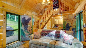 A cozy cabin interior with a wooden ceiling, a sofa with a blanket, and a staircase leading to an upper level. - Cozy Log Cabin on 11 Acres: 3 Mi to Cherokee Lake! (Bean Station)