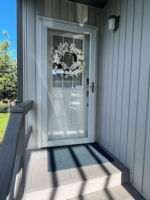 A white door with a frosted glass panel, a white wreath, and a doormat on a wooden porch.