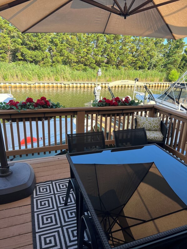 A deck with a table and chairs, overlooking a body of water with boats and greenery.