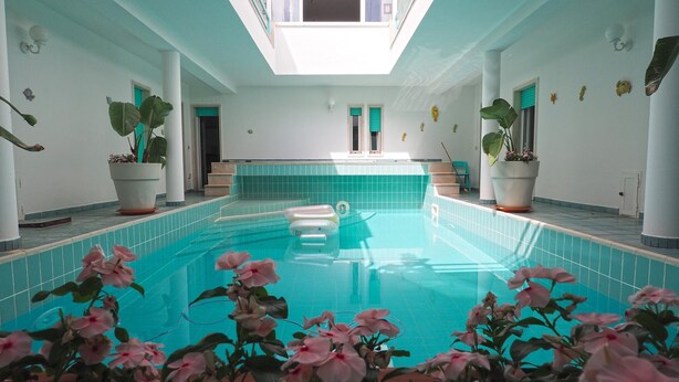 An indoor swimming pool with a clear blue tile floor, potted plants, and a view of the sky through the ceiling.