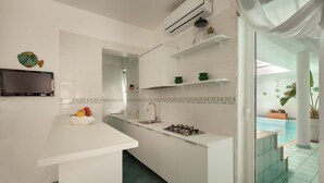 Eine moderne Küche mit weißen Schränken, einem eingebauten Gasherd, einem Spülbecken und einer Obstschale auf der Arbeitsplatte. An der Wand ist ein Flachbildfernseher angebracht und ein kleines Regal mit Dekorationen.