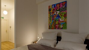 Una camera da letto con un letto, cuscini bianchi, un quadro astratto dai colori vivaci sulla parete e un comodino con una lampada.