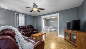A living room with a leather sofa, a wooden coffee table, a television on a wooden cabinet, and a ceiling fan. - Relaxing Home on the Outskirts! (Independence)