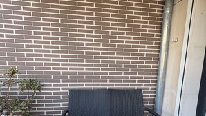 En mörk, flätad bänk placerad mot en tegelvägg, en krukväxt och ett träbord på en uteplats med kakelplattor.