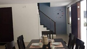 Comedor con una mesa rectangular dispuesta para seis personas, rodeada de sillas. Un jarrón grande se encuentra sobre la mesa, y hay una escalera que conduce a un nivel superior.