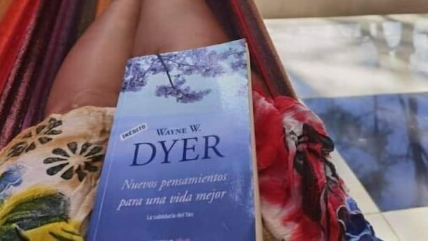 Una persona relajándose en una hamaca junto a una piscina, leyendo un libro titulado 'DYER' de Wayne W. Dyer.