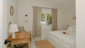 A bedroom with a bed, a wooden table with a lamp, a chair, a window with curtains, and a wall-mounted decorative item. - Casa Familia - House for 4 (Sa Pobla)