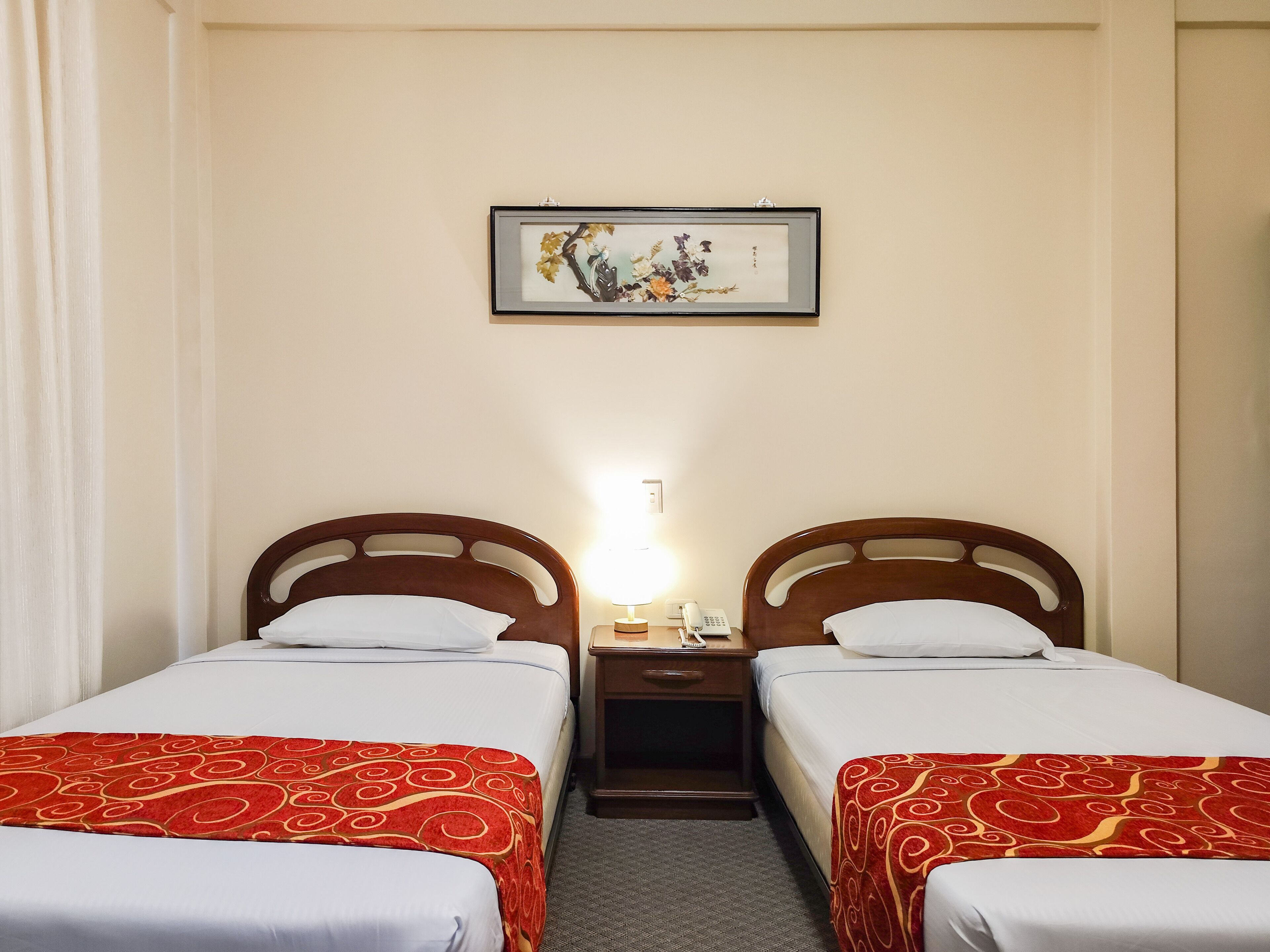 A hotel room with two single beds, each with a red patterned bedspread, a wooden headboard, and a bedside table with a lamp and a telephone.