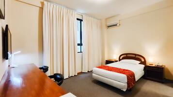 Quarto de hotel com cama, mesas de cabeceira, escrivaninha, cadeira, cortinas, ar condicionado e televisão.
