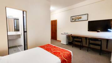 Quarto de hotel com cama, escrivaninha, cadeira, televisão e guarda-roupa.