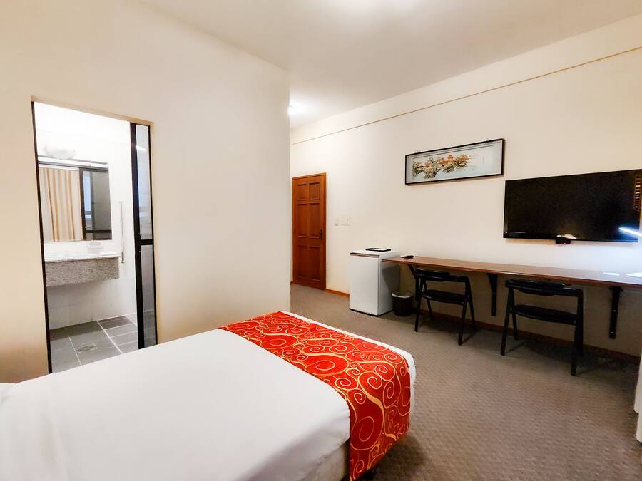 Quarto de hotel com cama, escrivaninha, cadeira, televisão e guarda-roupa.