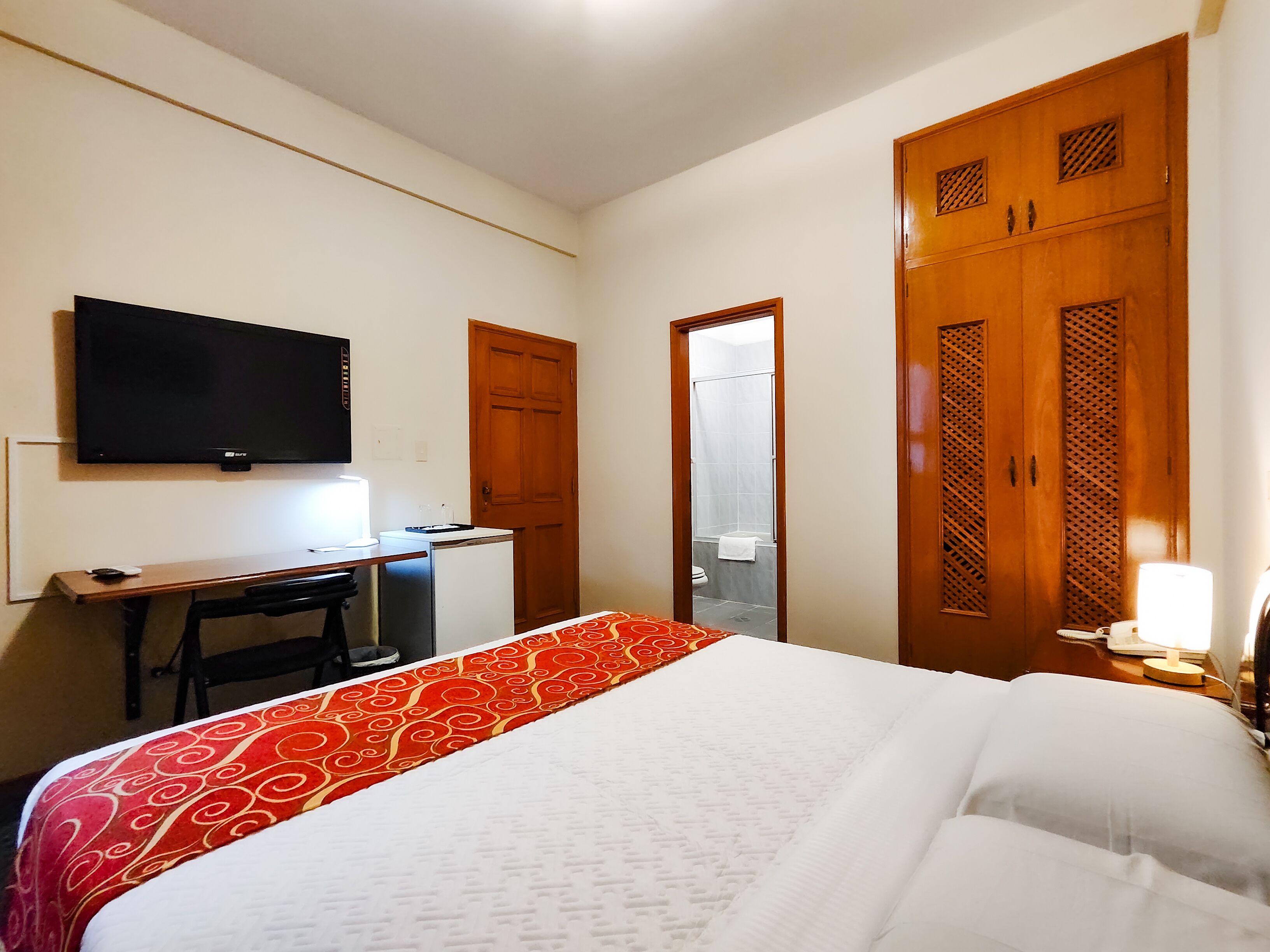 Una camera d'albergo con un letto, una scrivania, una televisione, un armadio e un bagno visibile attraverso una porta aperta.