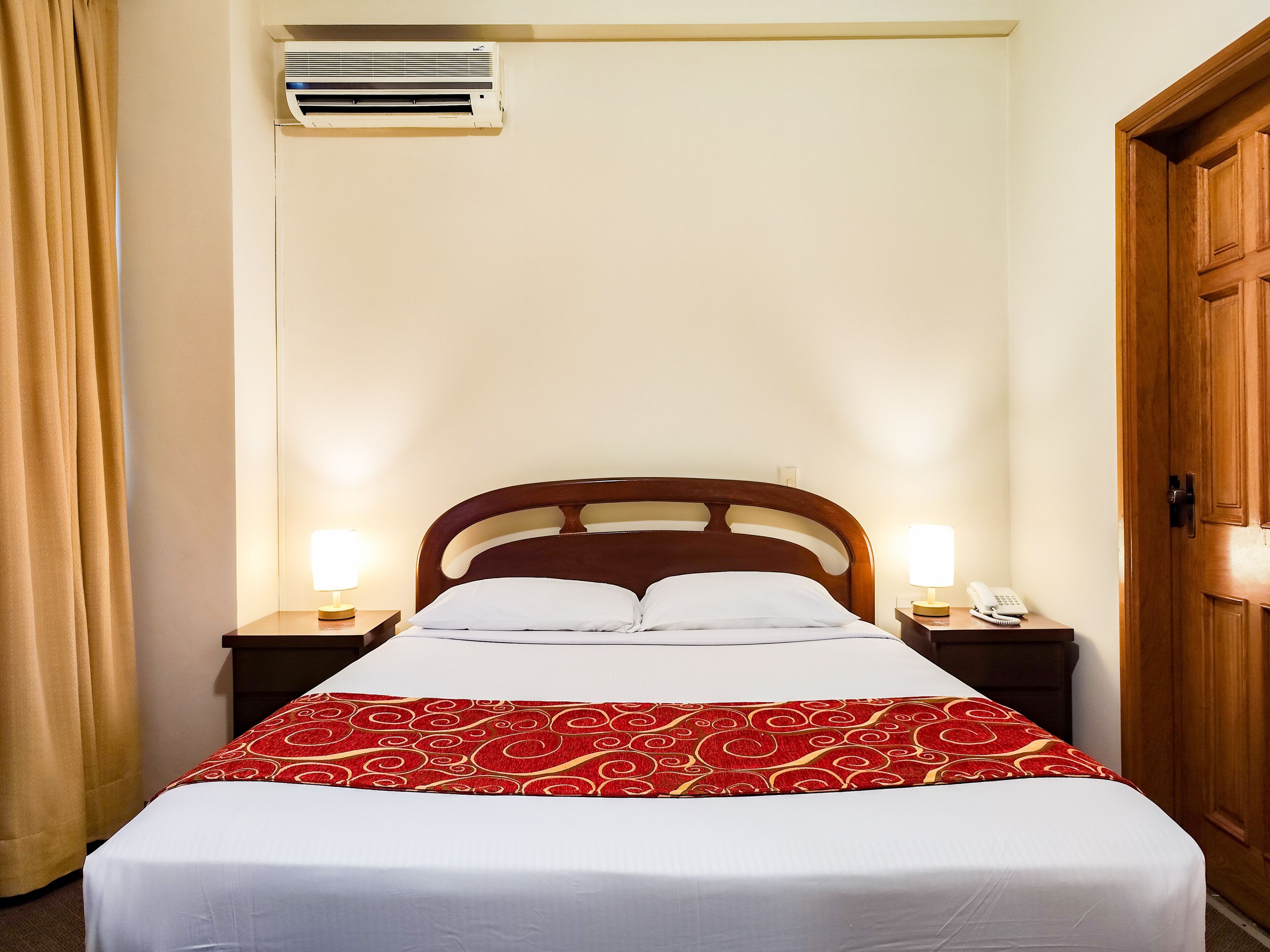 A hotel room with a bed, two bedside lamps, a telephone, and an air conditioning unit.
