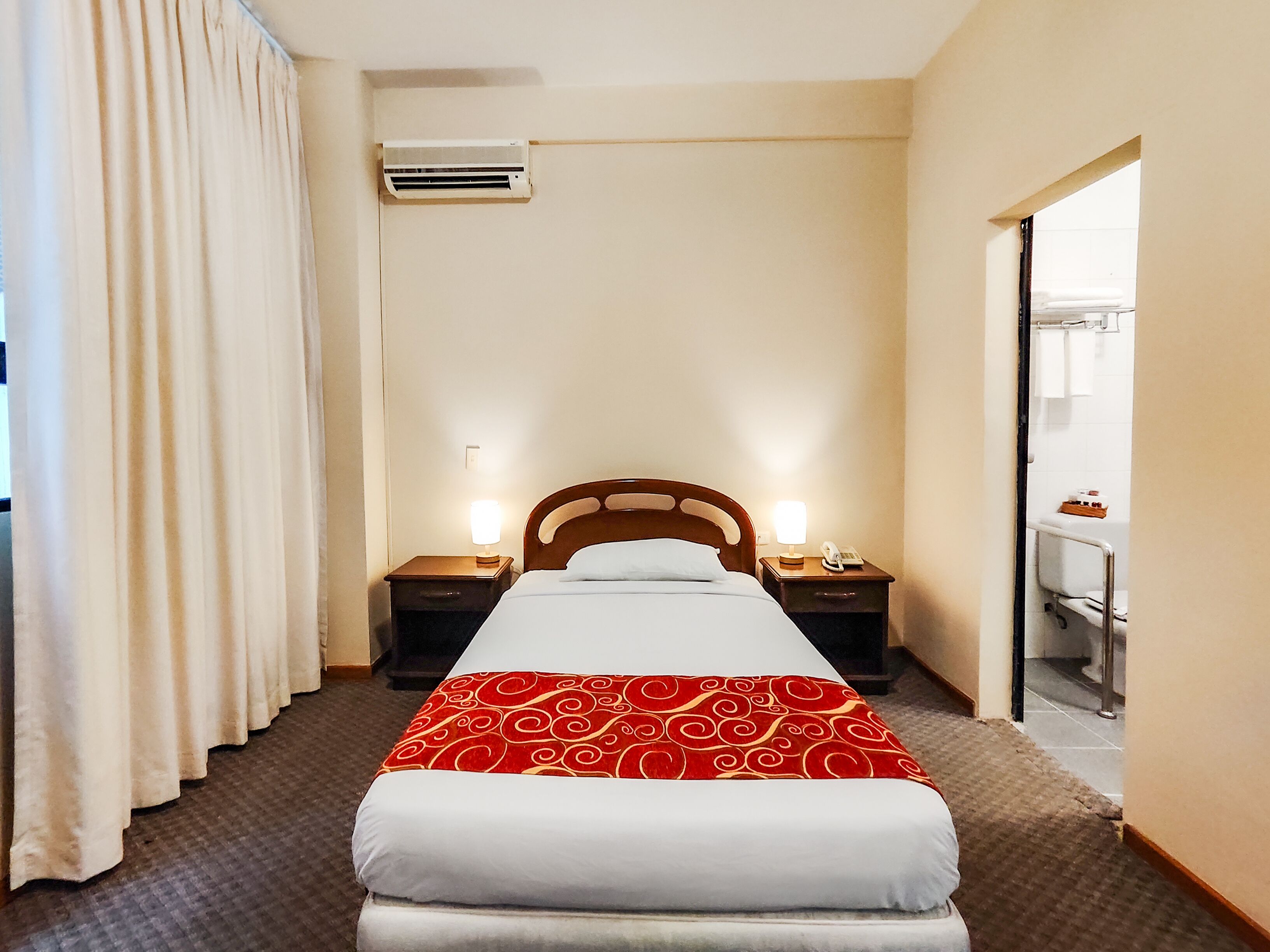 A hotel room with a bed, two bedside tables with lamps, an air conditioning unit, and a bathroom visible through an open door.