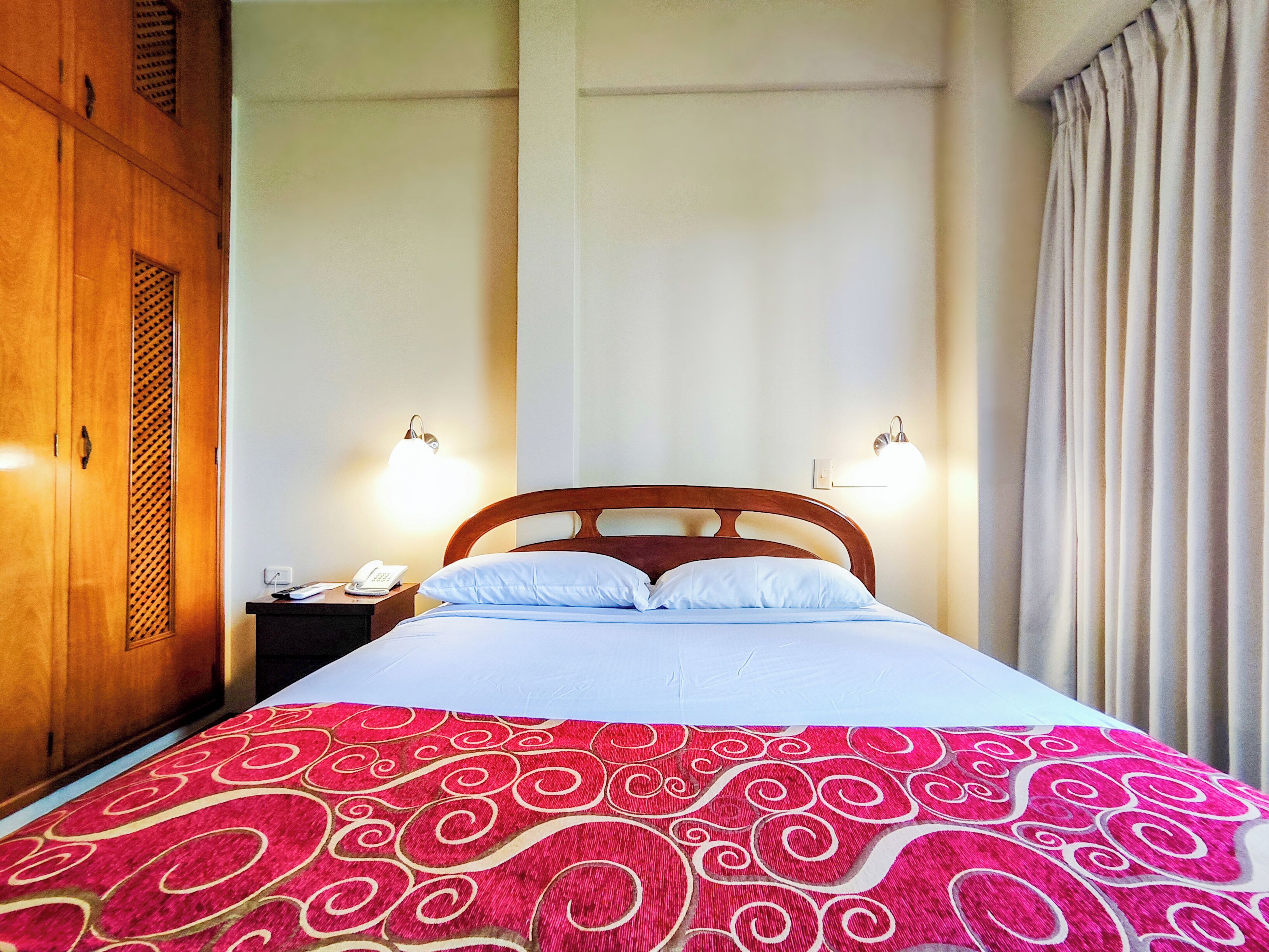 A hotel room with a bed, two bedside lamps, a nightstand with a phone, and a wooden wardrobe.
