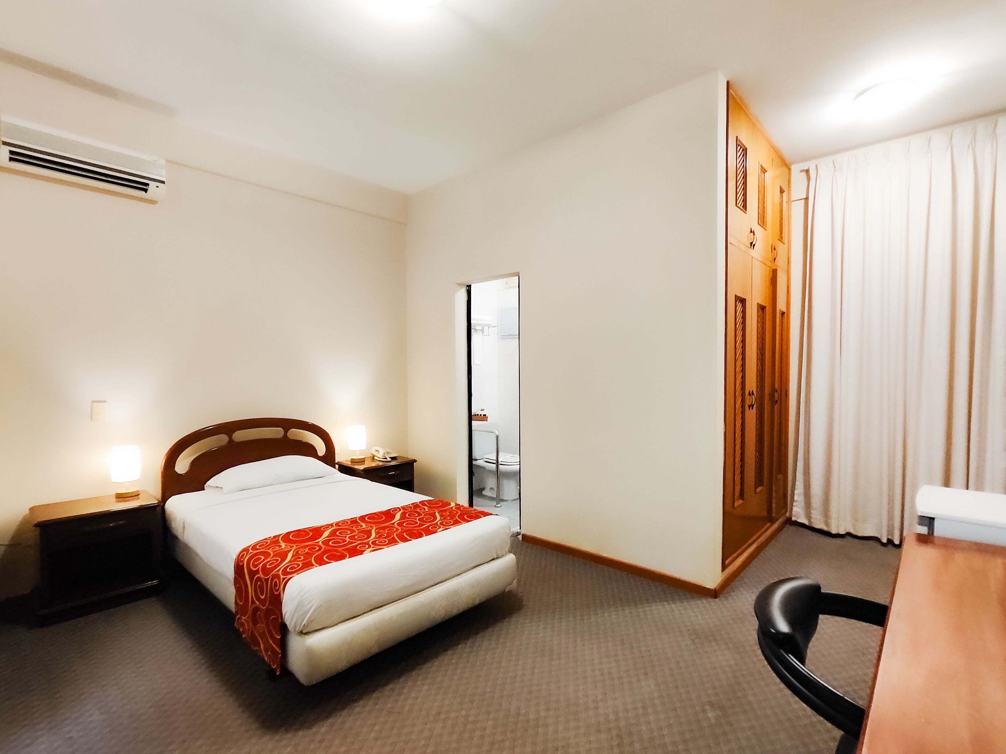 A hotel room with a bed, bedside tables, a desk, a chair, an air conditioning unit, and a door leading to a bathroom.
