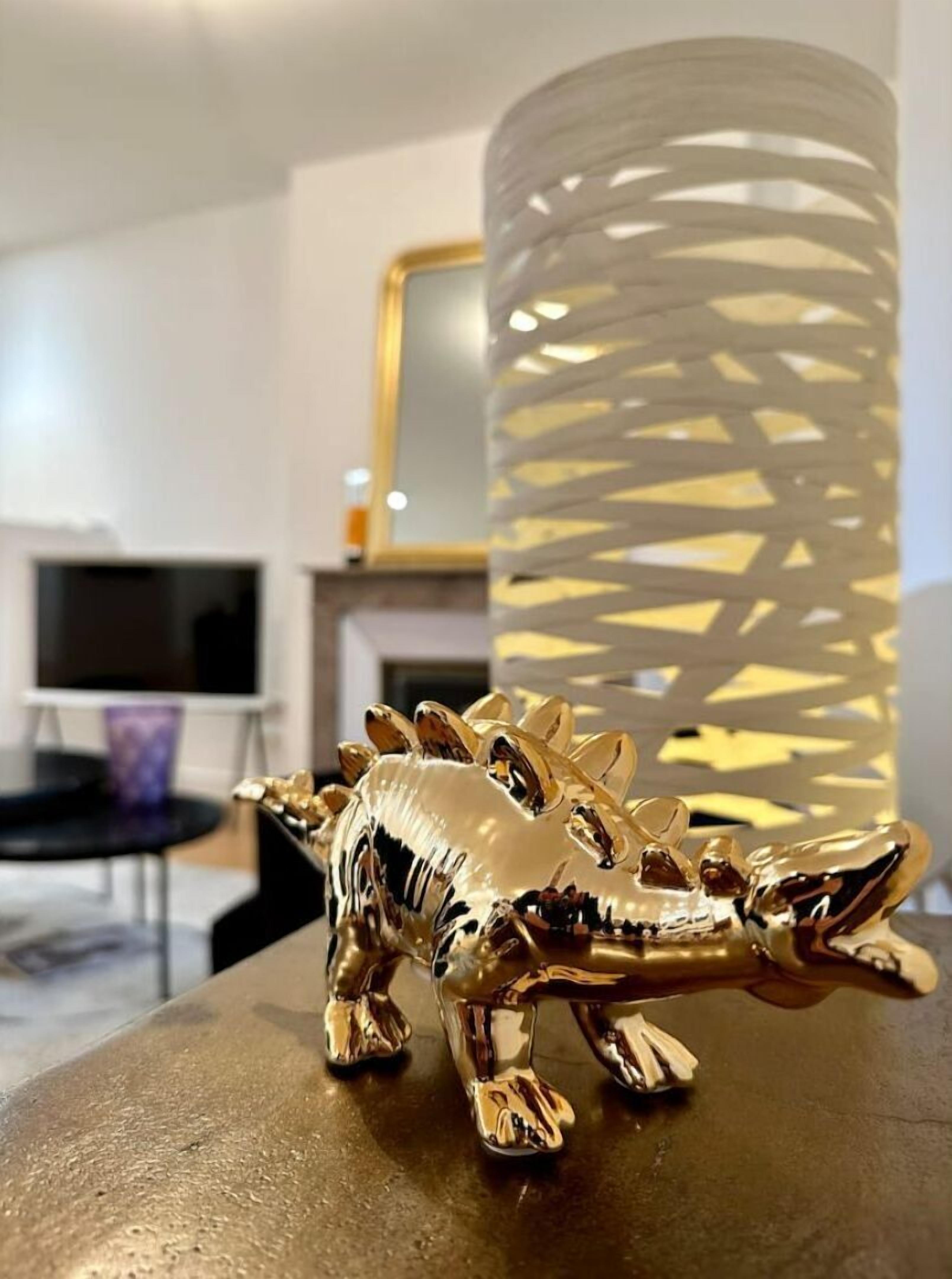 Une statuette de dinosaure dorée est posée sur une table ronde dans un salon moderne, avec un grand vase décoratif en arrière-plan.