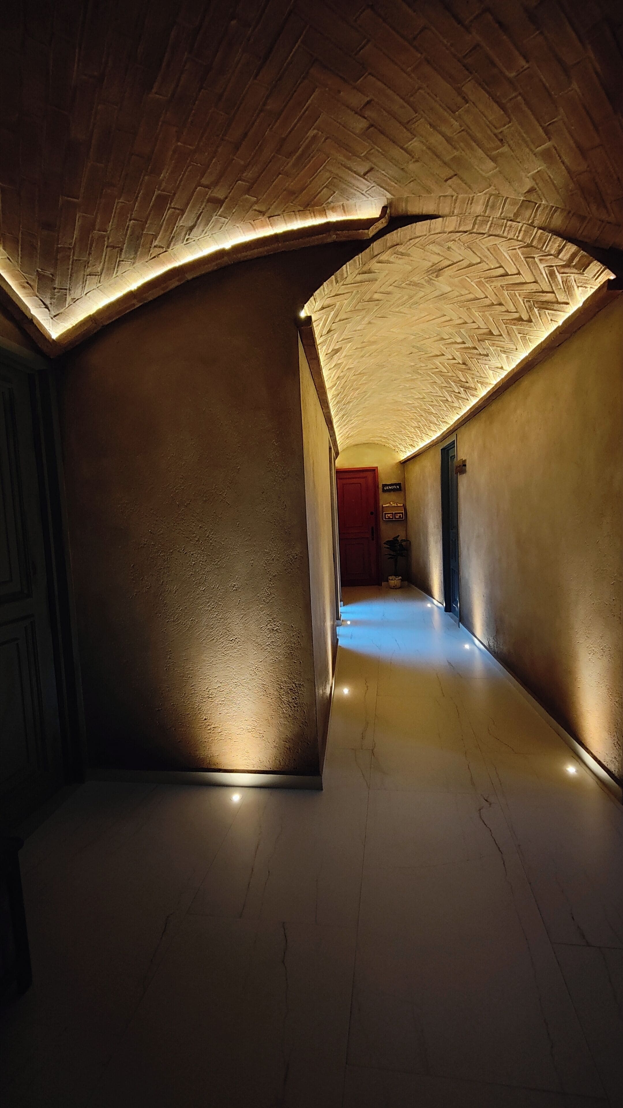 A corridor with a herringbone-patterned ceiling and a polished floor, leading to a door labeled "GENOVA".