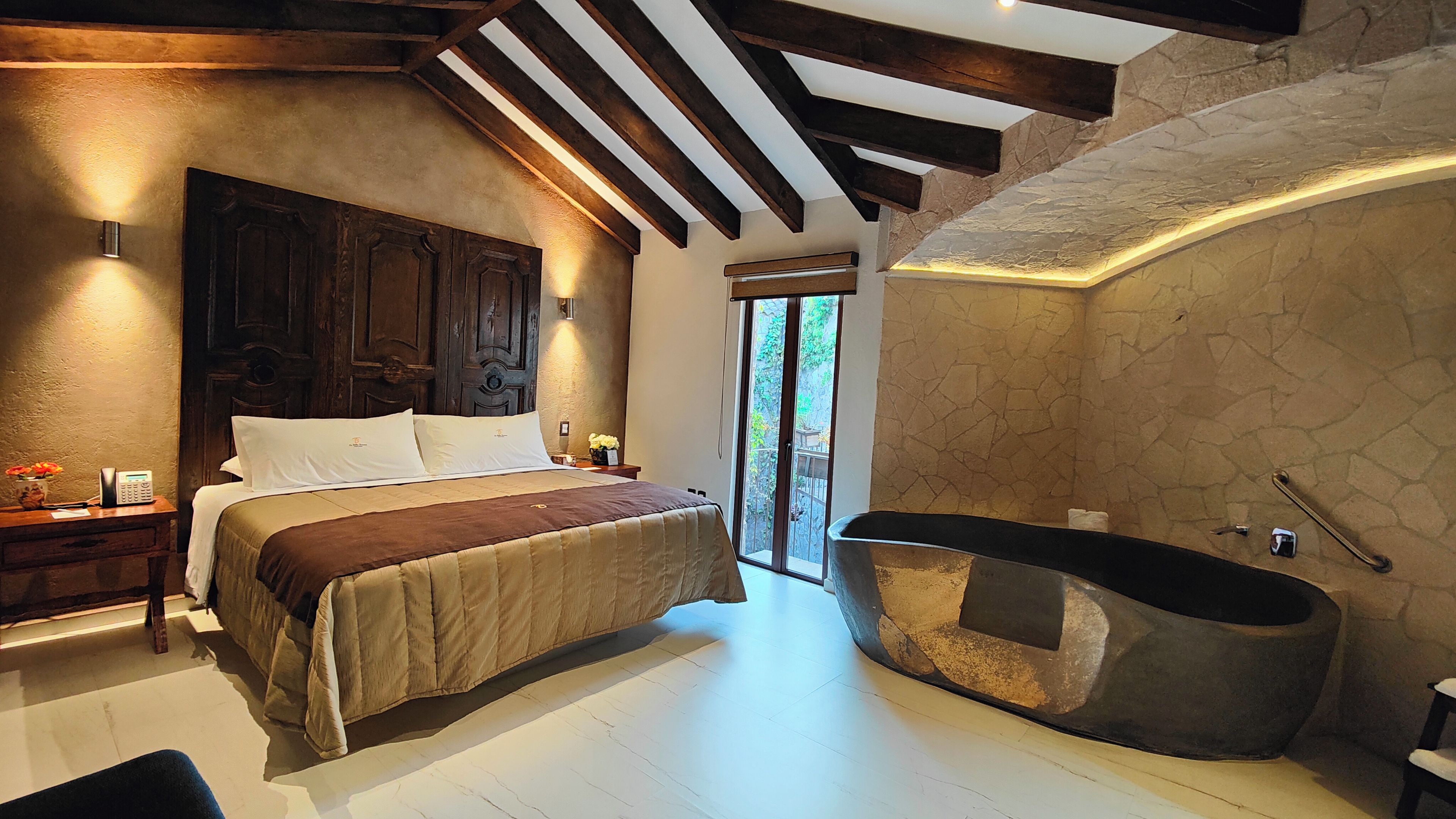 Un dormitorio con una cama grande, un cabecero de madera, una mesita de noche con un teléfono y un florero, y un baño con una bañera.