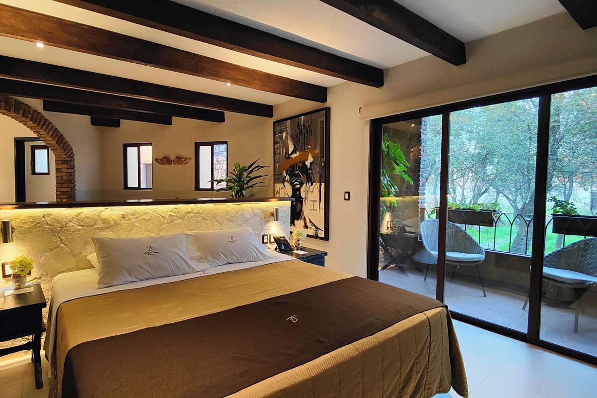 A modern bedroom with a large bed, a stone archway, a bathroom with a glass door, and a balcony with outdoor seating.
