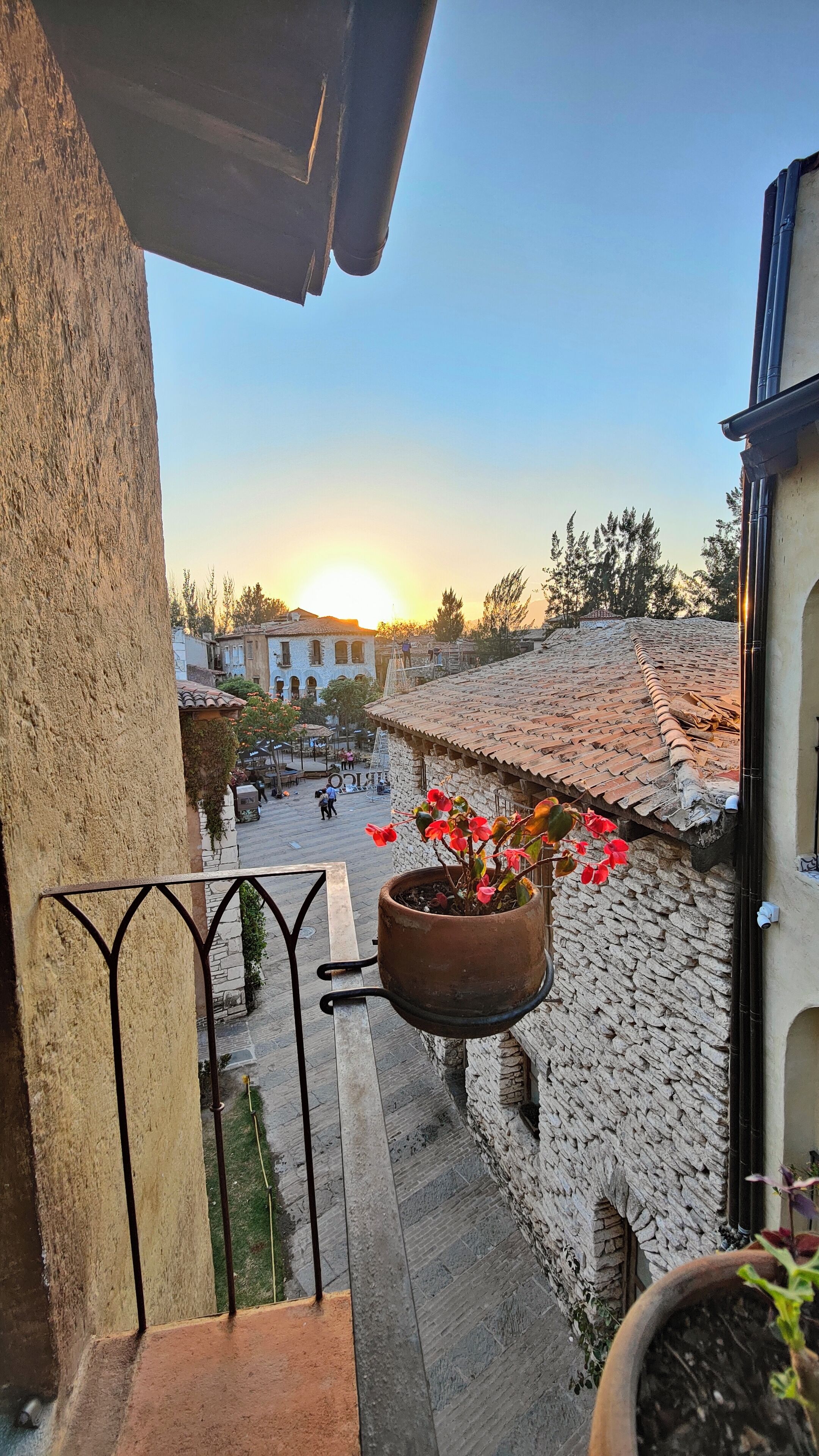 A balcony with a hanging flower pot and a view of a town square during sunset.