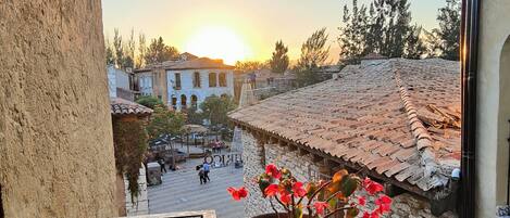 A balcony with a hanging flower pot and a view of a town square during sunset.