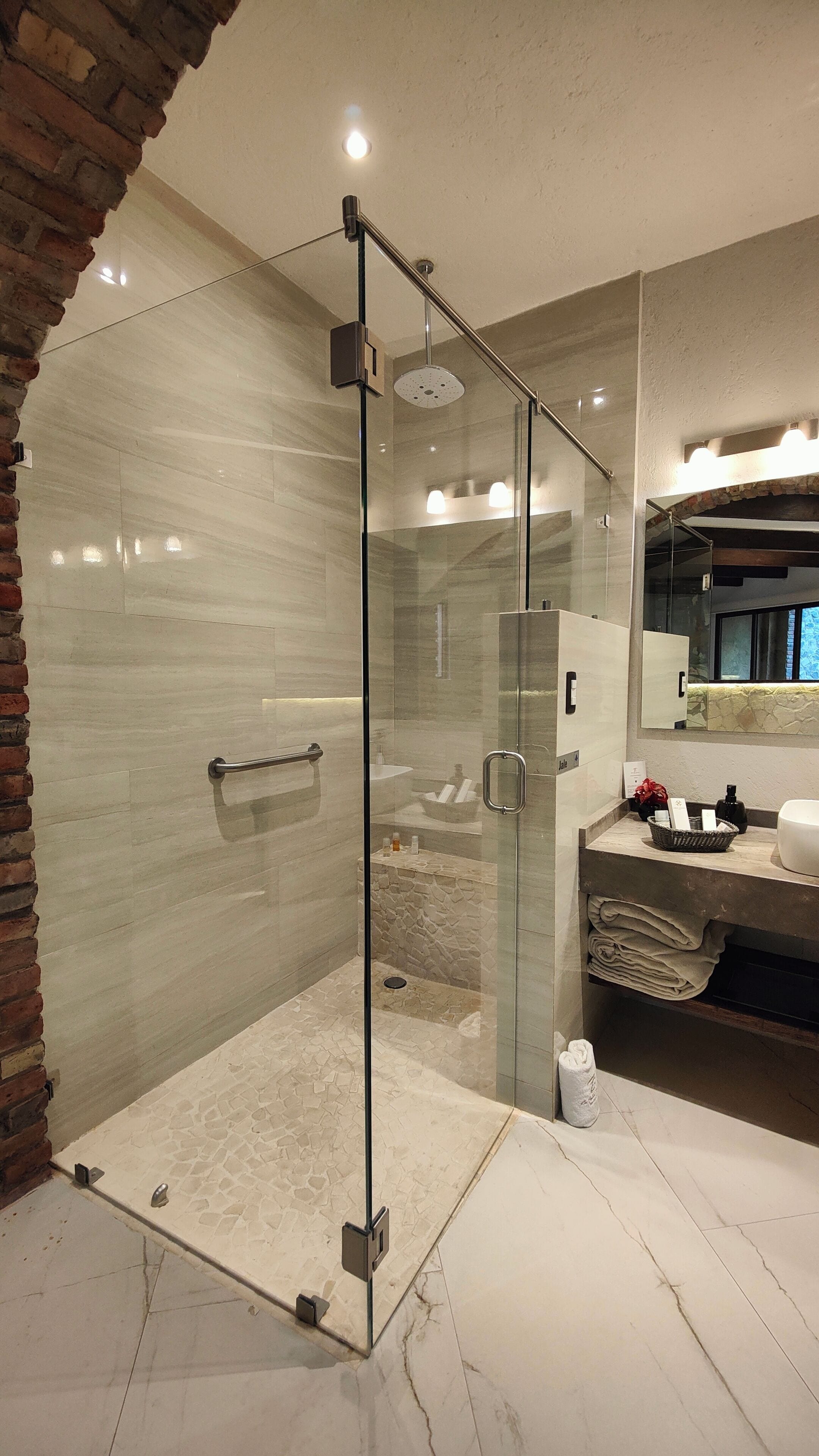 A modern bathroom with a glass-enclosed shower, a marble-tiled floor, and a visible sink area with toiletries.