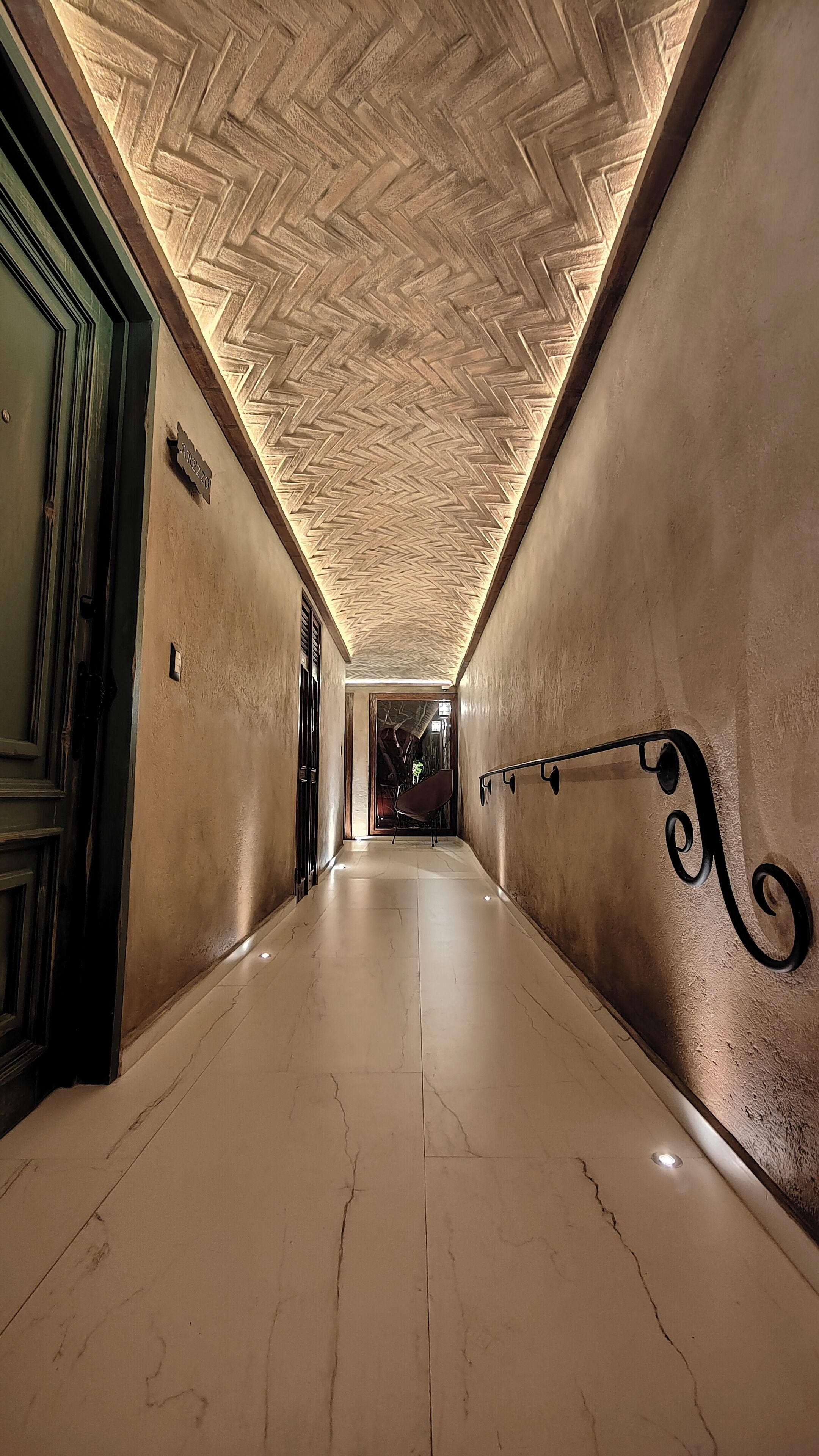 Un couloir avec un plafond orné d’un motif en chevrons, un sol en marbre et une balustrade en métal.