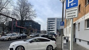 Un espace de stationnement avec des voitures, un bâtiment à l’architecture moderne et un panneau indiquant un espace réservé au stationnement des véhicules électriques.