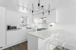 Une cuisine moderne avec des meubles blancs, un îlot central, un luminaire suspendu et une fenêtre donnant sur des immeubles.