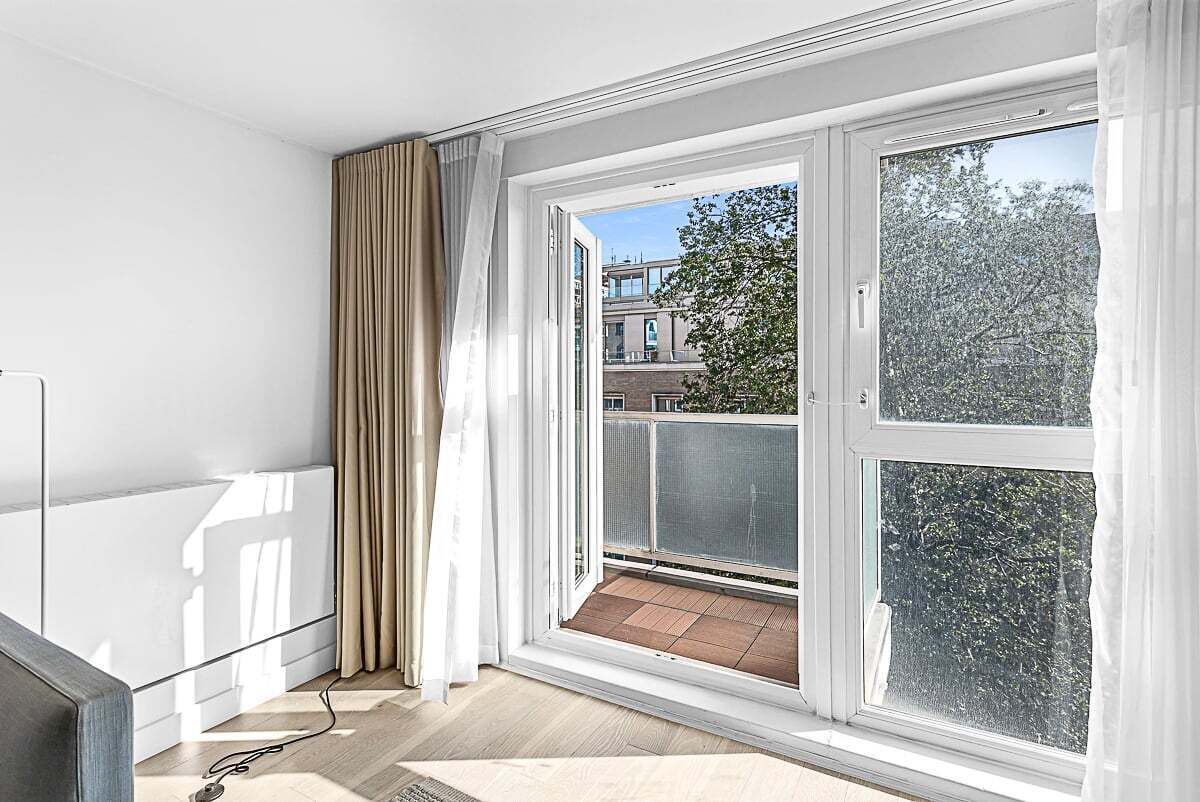 A room with a large window, a balcony, and a view of buildings and trees.