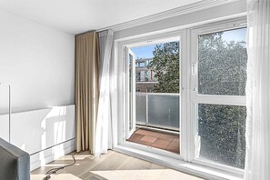 Una habitación con un ventanal, un balcón y vistas a edificios y árboles.