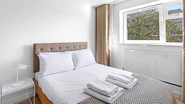 A hotel room with a bed, a nightstand, a window with curtains, and a view of trees outside. - Belgravia Base: 2BR Near Victoria/Sloane (London)