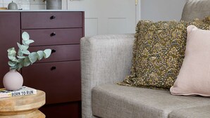 Een moderne woonkamer met een grijze bank, een kussen met een patroon, een houten bijzettafeltje met een vaas, en een donkerbruine kast met lades.
