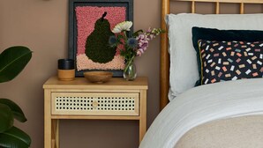 Een slaapkamer met een houten hoofdbord, een nachtkastje met een kunstwerk in een lijst, een vaas met bloemen en een grote plant in pot.