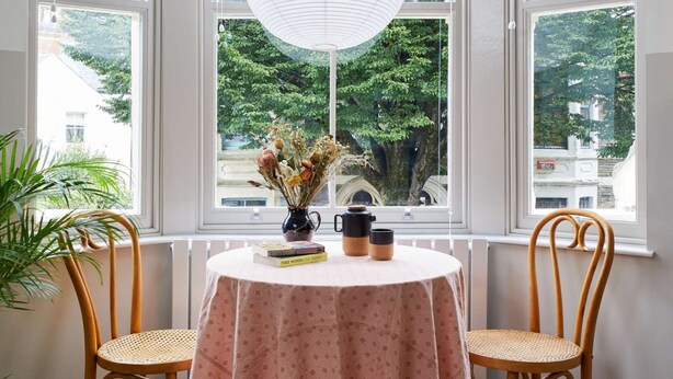 Een erker met witte blinds, een ronde tafel met een roze tafelkleed, twee houten stoelen en een hangende witte hanglamp.