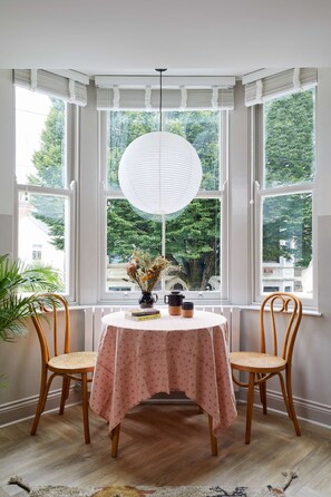 Une fenêtre en saillie avec des stores blancs, une table ronde recouverte d’une nappe rose, deux chaises en bois et une suspension blanche.